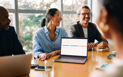 Business professionals share a laugh during a meeting, engaging over a laptop with charts displayed.